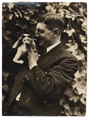 Photo of man in suit and tie looking at a cat, held affectionately on his shoulder, standing near foliage.