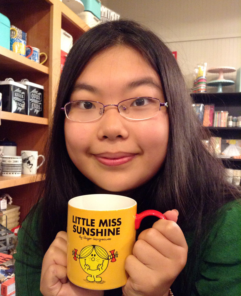 A photo of a woman with long black hair and glass smiling while holding a little miss sunshine mug