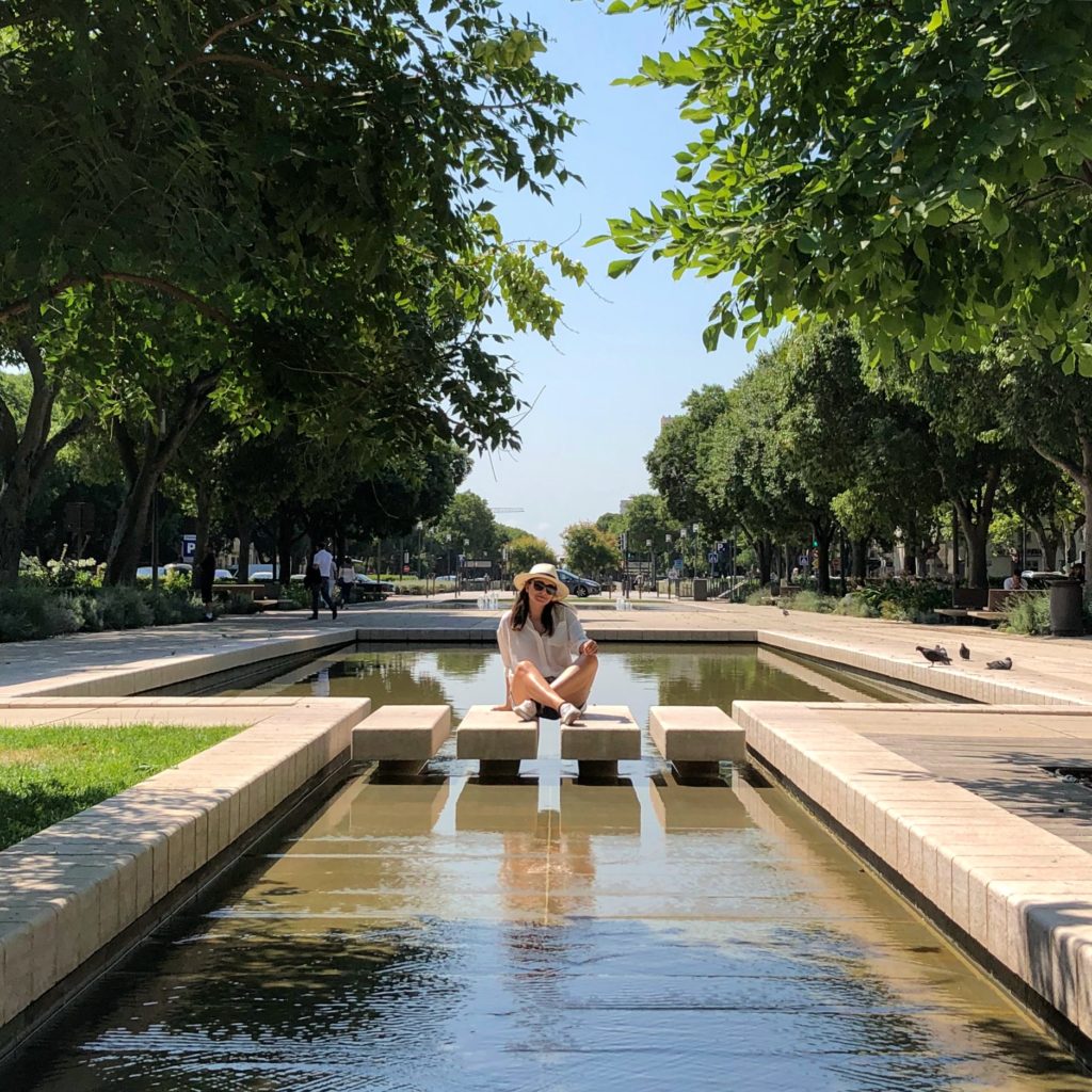 A photo of trees on either side with a woman sitting in the center surrounded by a fountain