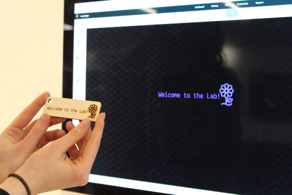 Hands holding a small wooden sign that reads, "Welcome to the Lab!" in front of a computer screen.