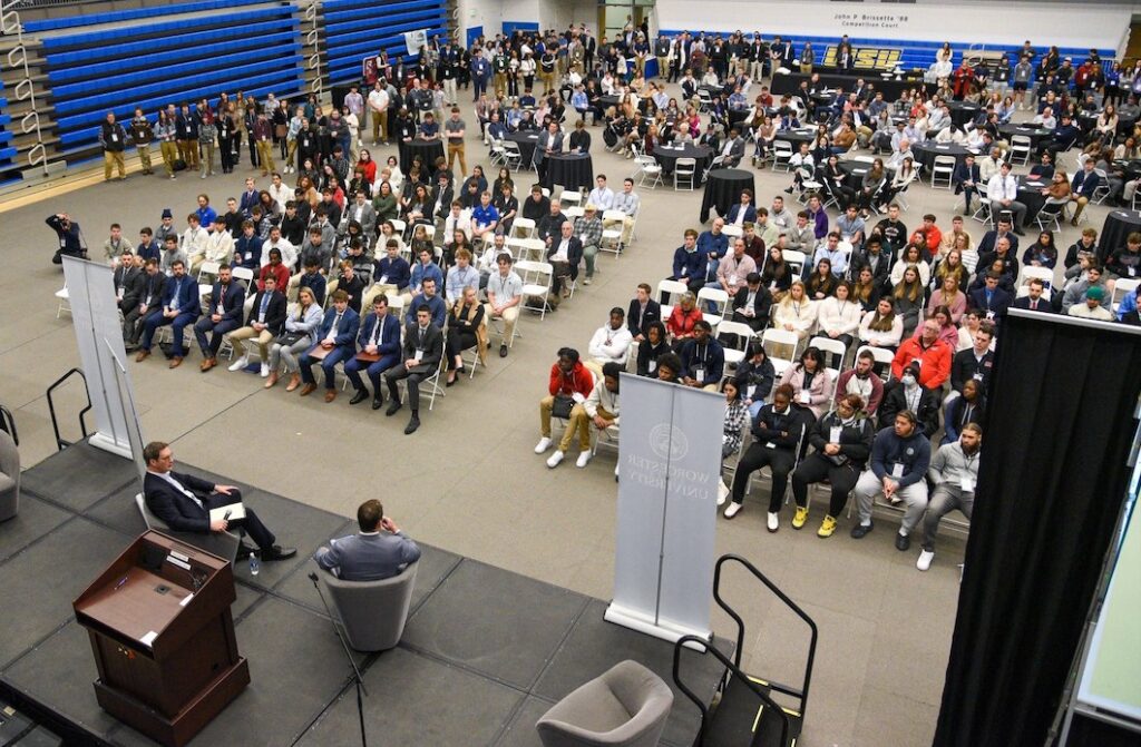 A gymnasium full of students in chairs listenting to two men on stage talk about sports. 