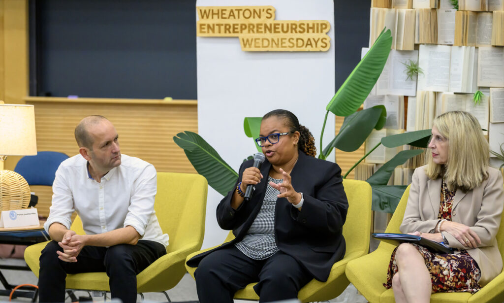 Three individuals sit in chairs while the middle person speaks with a microphone.