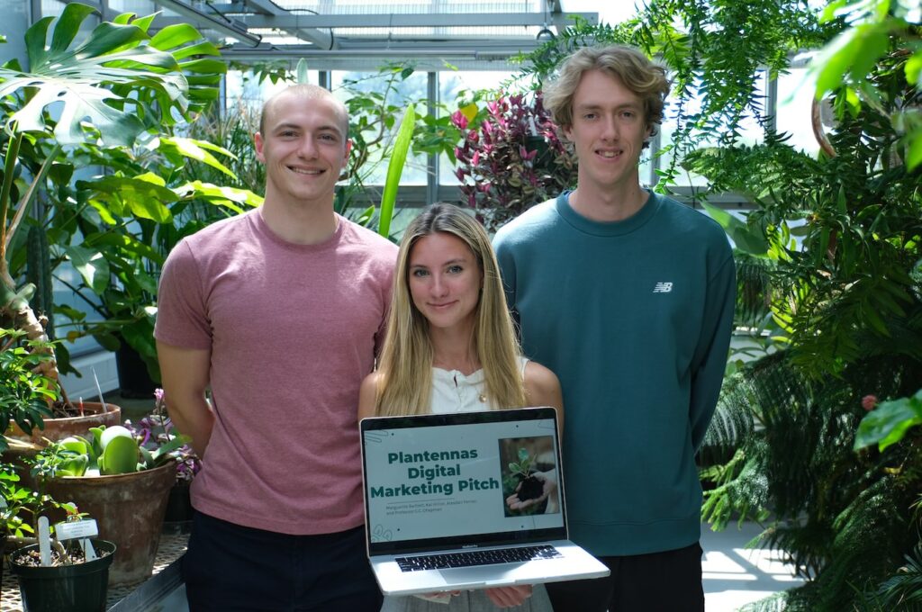 Three college students stand in the middle of a greenhouse holding a laptop that reads Plantennas Digital Marketing Pitch on the screen.