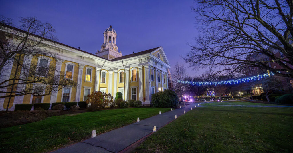A yellow building is illuminated at night, with a lighted walkway and strings of lights on the right side of it.