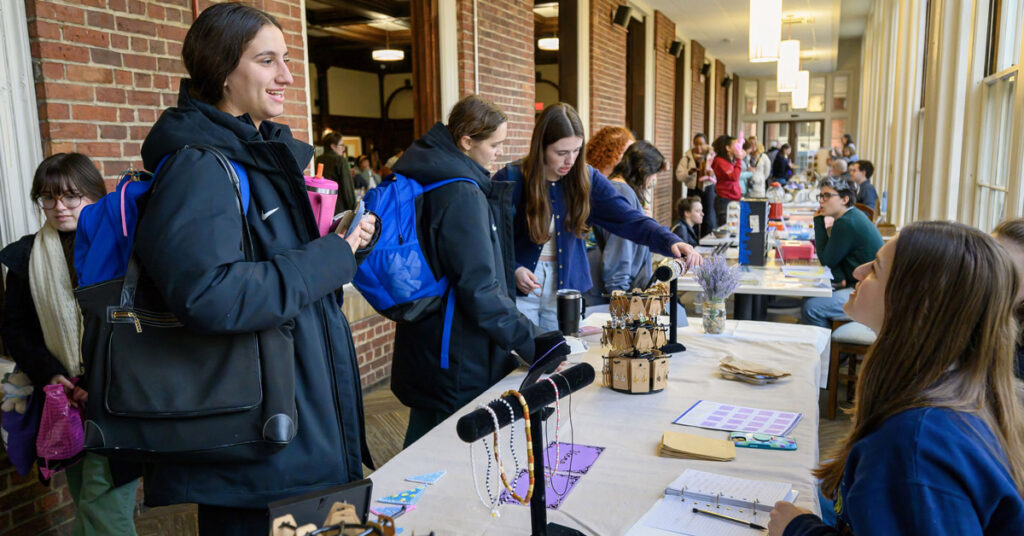 A smiling woman with dark hair wearing a dark winter coat speaks with a woman sitting behind a table. A row of tables with others speaking similarly are visible in the background.