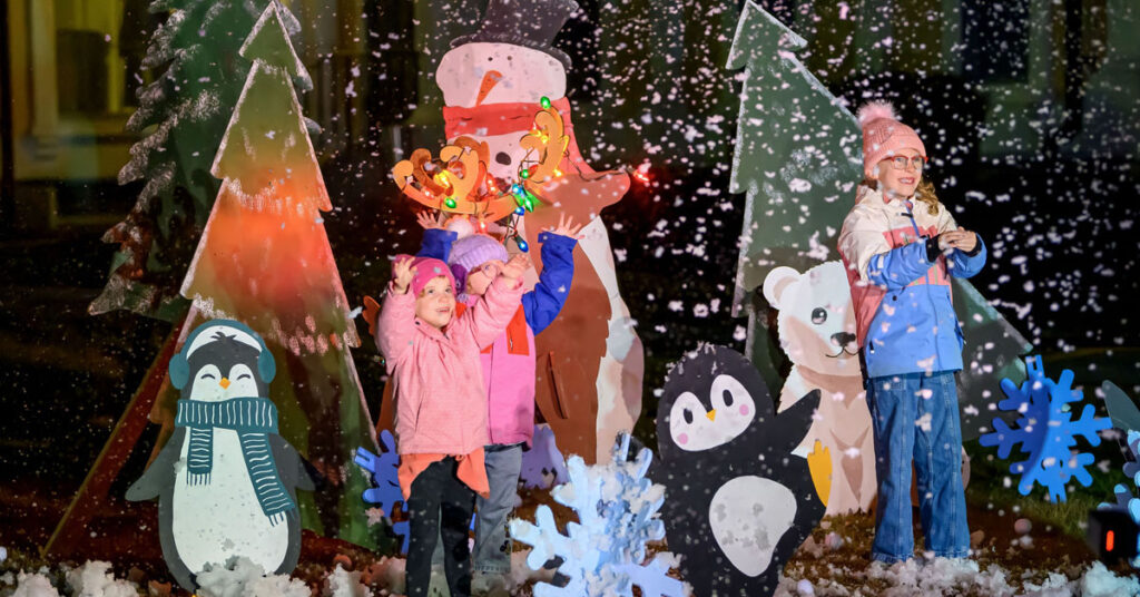 A few smiling young girls wearing winter clothing stand outside, surrounded by cutouts of animals, christmas trees and snowflakes.