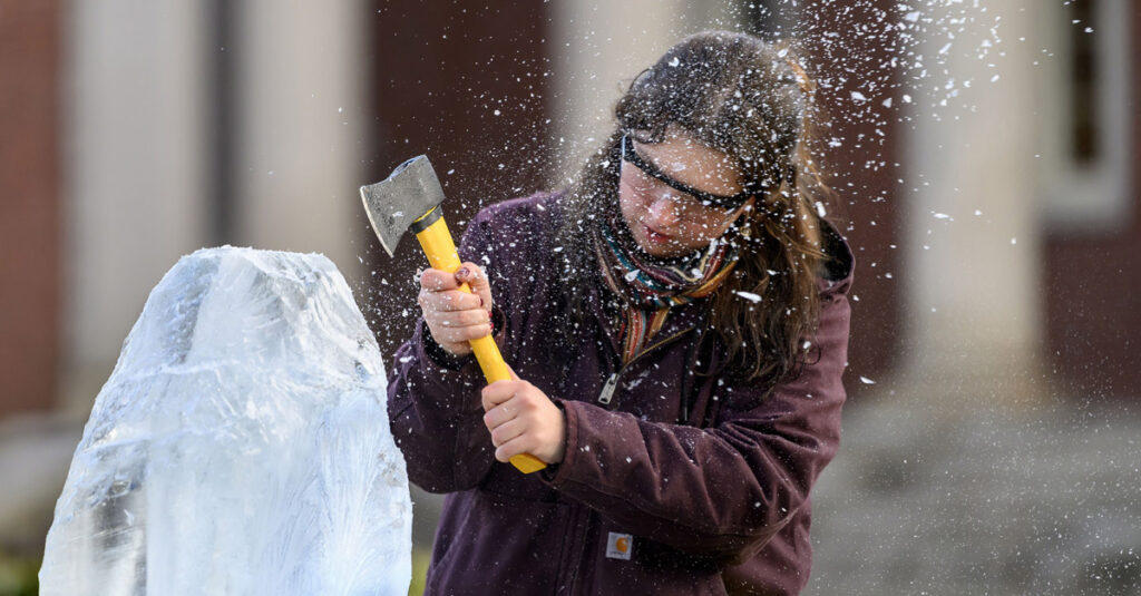 An individual with long dark hair wearing a dark winter coat and safety glasses wield a small axe, chipping at a block of ice.