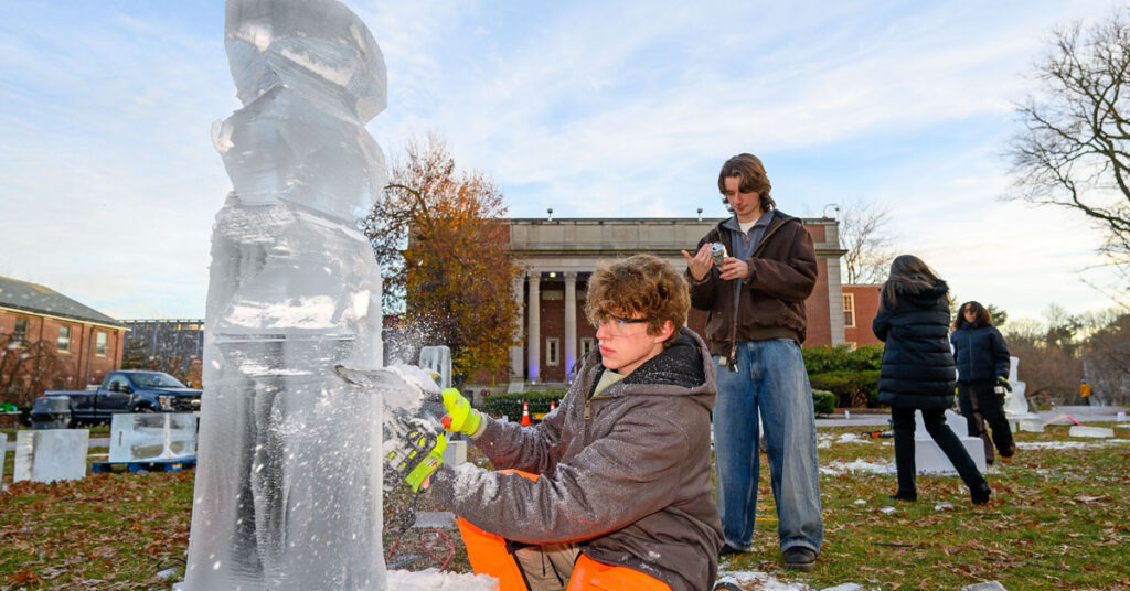 A young man wearing winter clothing and safety classes carves a block of ice with a chain saw while a young man watches from behind.