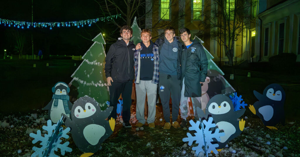 A group of four smiling young men wearing winter clothing stand outside, surrounded by cutouts of animals, christmas trees and snowflakes.