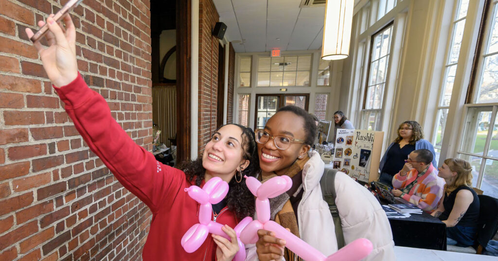 Two women smile while holding balloon animals and taking a selfie photo.