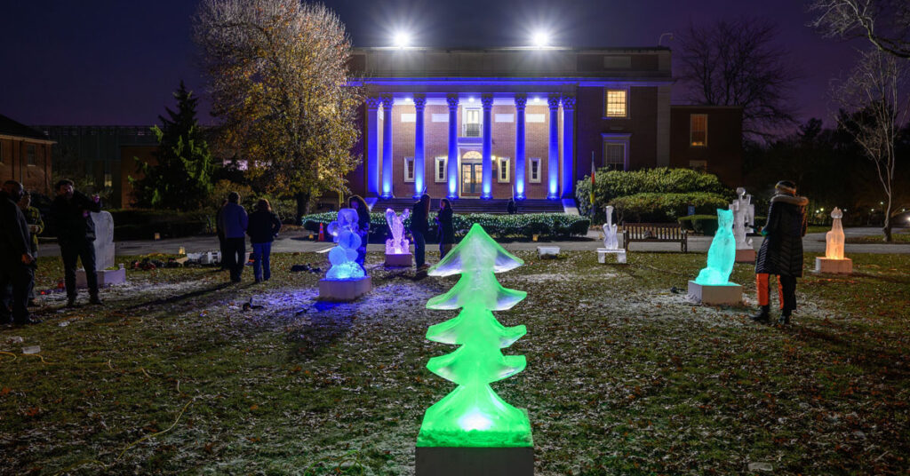 An illuminated green ice sculpture, in the shape of a pine tree, is the centerpiece of the image. Several other illuminated ice sculptures are visible in the background in front of a building with large columns and lights on at its entrance