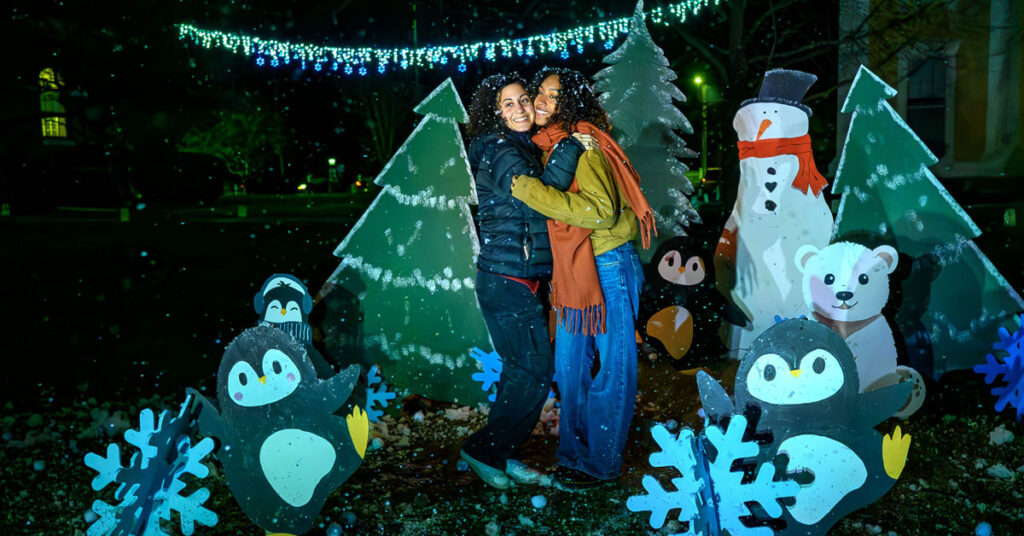 Two smiling women embrace while wearing winter clothing outside, surrounded by cutouts of animals, christmas trees and snowflakes.