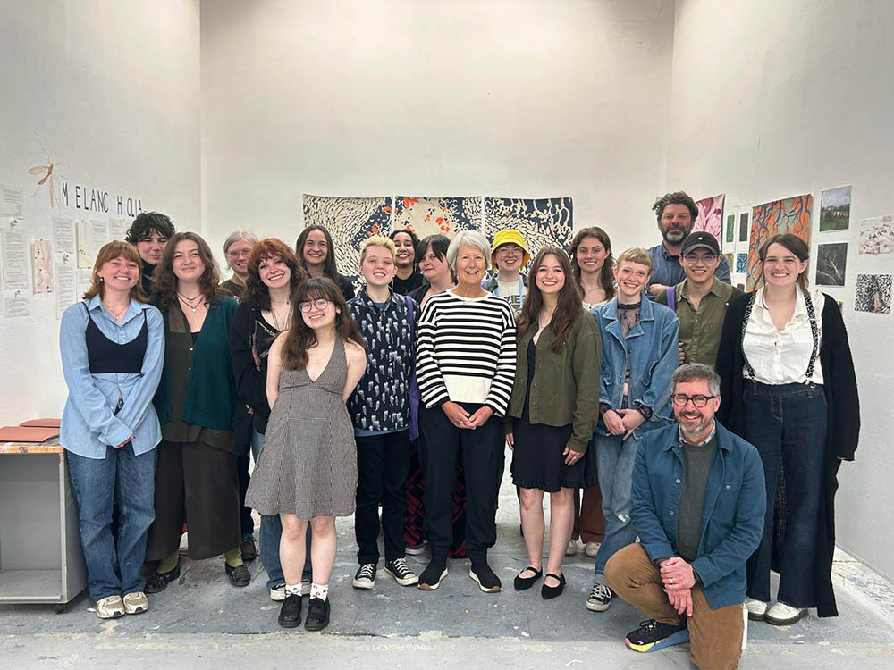 A formal group portrait of 19 people in a gallery. 16 students who participated in the Arts in Ireland program. Professors Kelly Goff and Kent Shaw. Artwork can be seen behind them. 