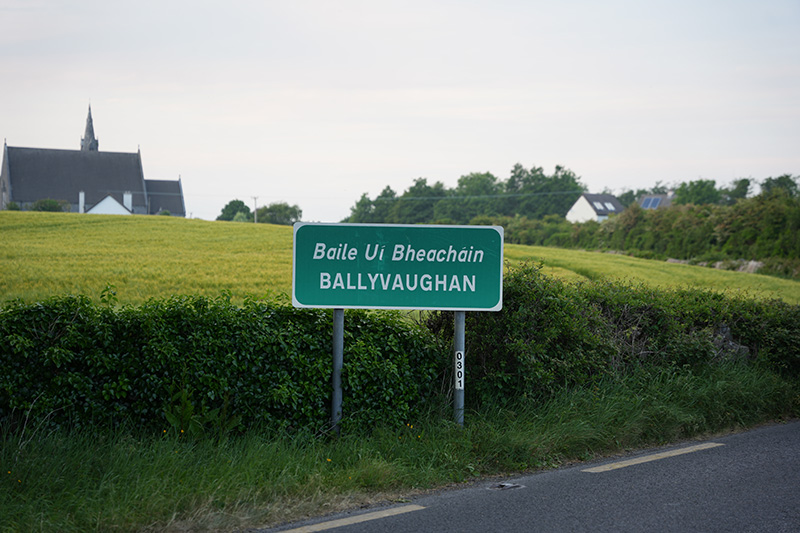 A portrait of the town sign for Ballyvaughan, Ireland that is next to a road. A bucolic field is behind the sign.