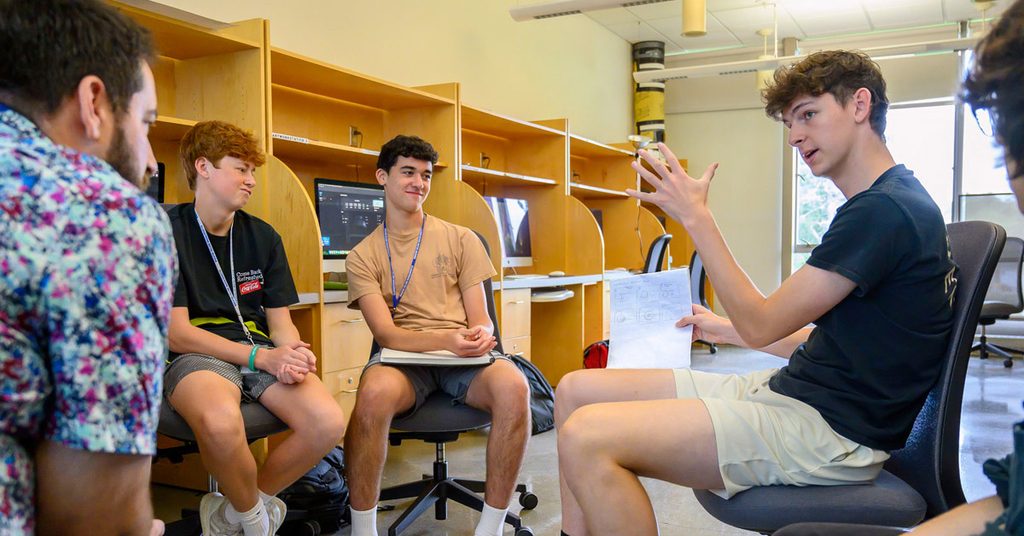 Three college students wearing shorts and T-shirts sit on chairs in a semi-circle in a classroom while speaking with an older gentleman with a beard and a patterned floral shirt, most likely a professor.