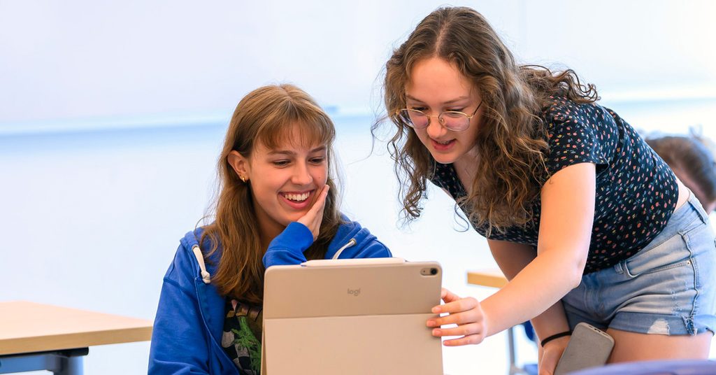 A young female with long brown hair smiles while looking at a computer screen. An older female with long brown hair wearing glasses, and floral patterned dark shirt and jeans stands beside her, holding the side of the computer screen with one hand while also looking at the screen.