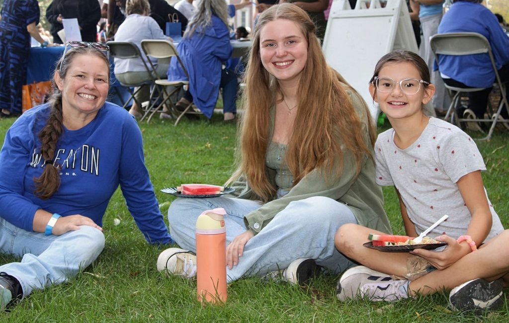 Six smiling females, including a child, are dressed casually and sit on grass while holding food on plates.