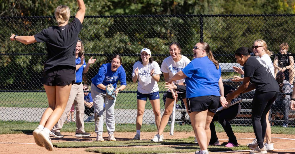 A group of excited women gather around home plate at a softball game as another woman jumps as she approaches the group.