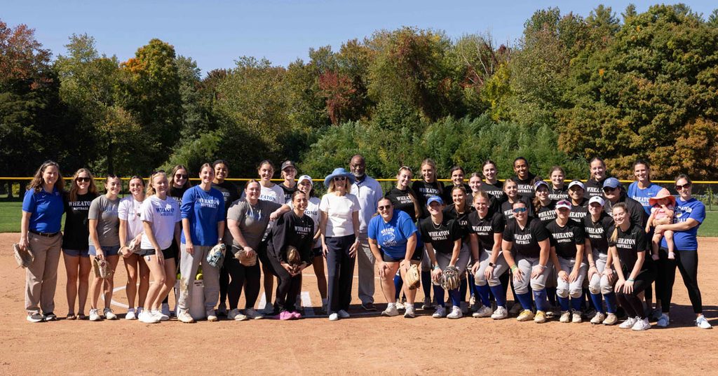A large group of women, many wearing softball gear, gather for a group photo on a softball infield.