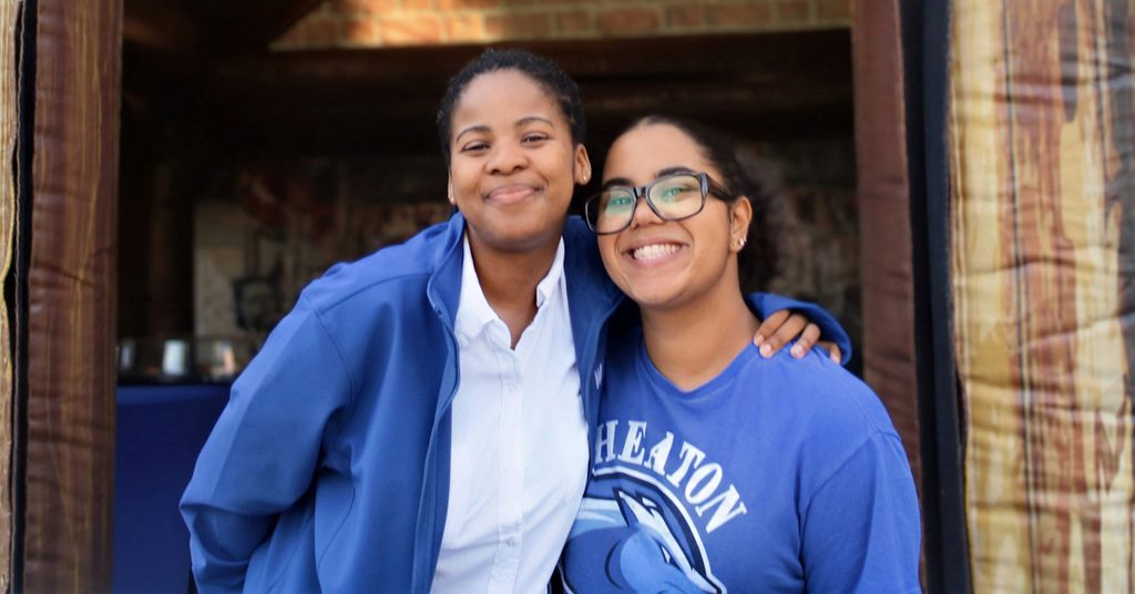 Two smiling women, with their arms around each other's back.