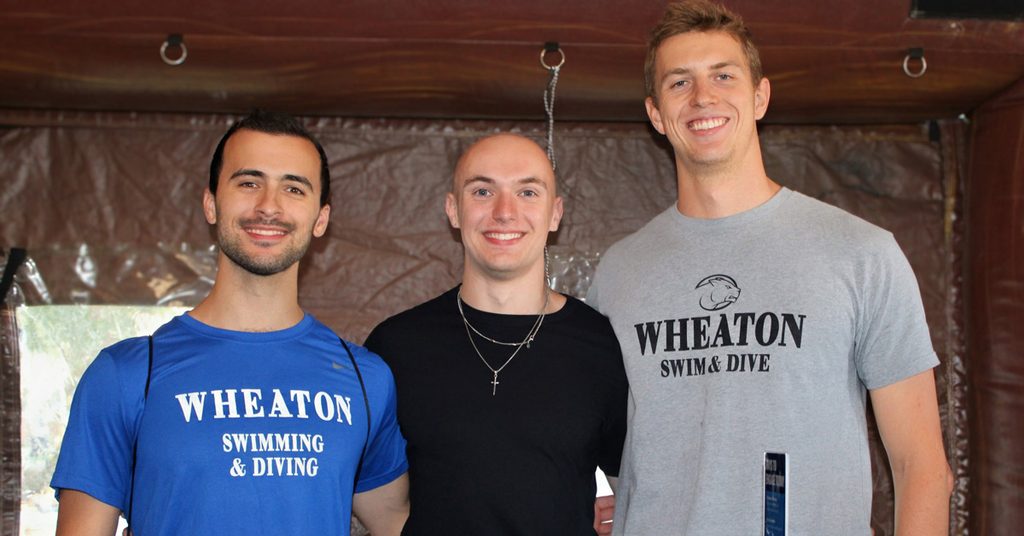 Three smiling young men each wear a T-shirt. Two of the T-shirts read Wheaton swimming & diving.