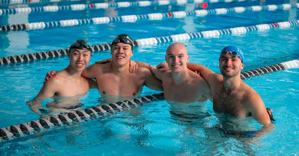 Four smiling men wearing swimming caps stand in a pool with arms around each other's back.