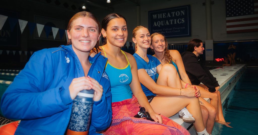 Four women wearing swim gear smile while sitting on a beach outside an indoor pool.