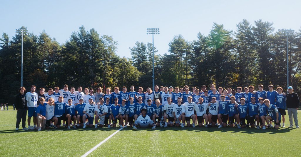 A large group of men, most wearing either white or blue uniforms, gather on an outdoor turf field for a photo.