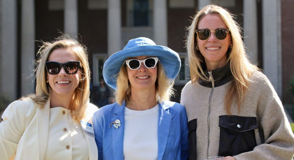 Three smiling women, each wearing sunglasses and a sweater, stand outside. A building with columns is in the background.