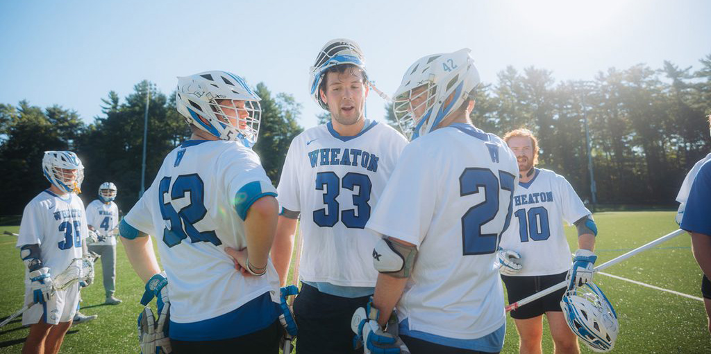 A group of three male lacrosse players, each wearing a white uniform with blue numbers and "Wheaton" written on the front, talk on a field. Other players in uniform are behind them.