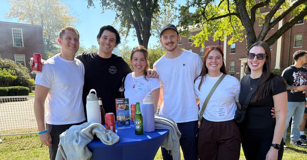 Six smiling young men and women, dressed casually, stand outside in front of a small round table adorned with beverages. Buildings and trees with green leaves are visible behind them.