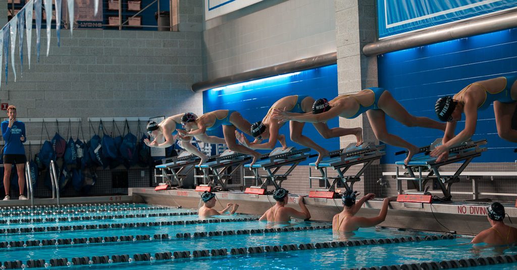 A group of five individuals wearing swimsuits and bathing caps dive into an indoor pool to start a race.