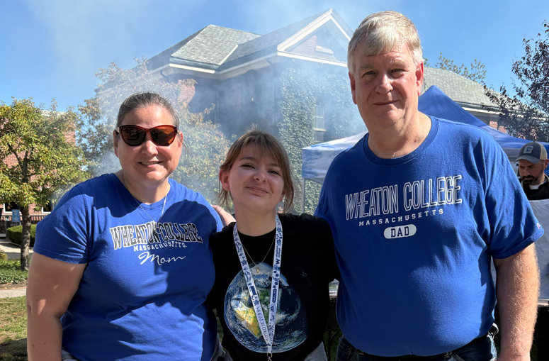 A young individual wearing a black T-shirt, stands between an older woman and man, both wearing blue T-shirts that read Wheaton College. Buildings and trees with green leaves are visible behind them.
