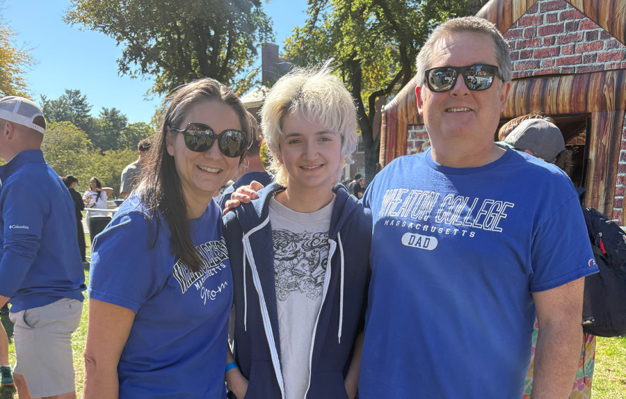 A young individual wearing a dark sweatshirt over a light T-shirt, stands between an older woman and man, both wearing blue T-shirts that read Wheaton College. Buildings and trees with green leaves are visible behind them.